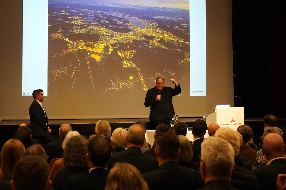Ein Mann im dunklen Anzug steht auf einer Bühne und referiert. Im Hintergrund ist ein Luftbild des Grossraums Zürich bei Nacht zu sehen.